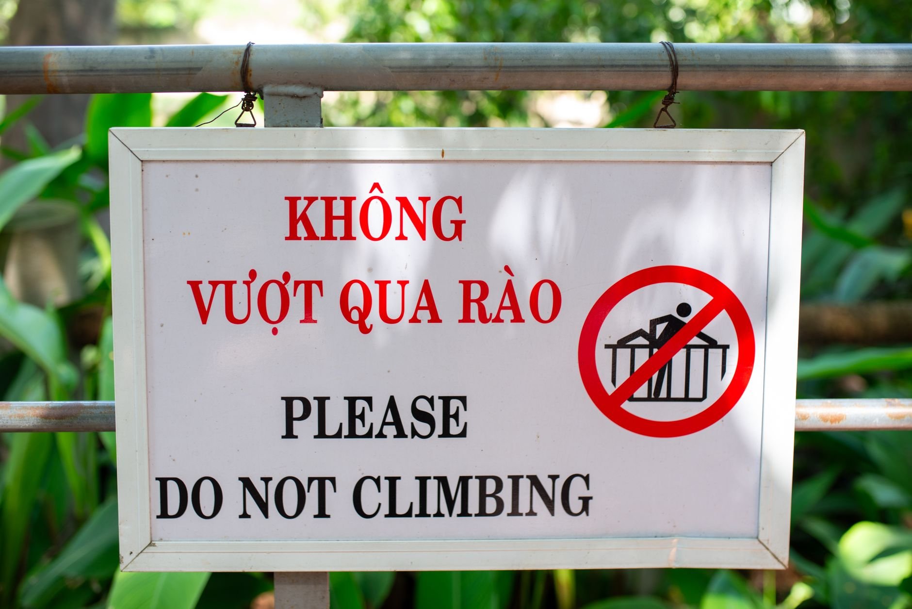 Bilingual safety sign warning against climbing, displayed outdoors with green foliage.